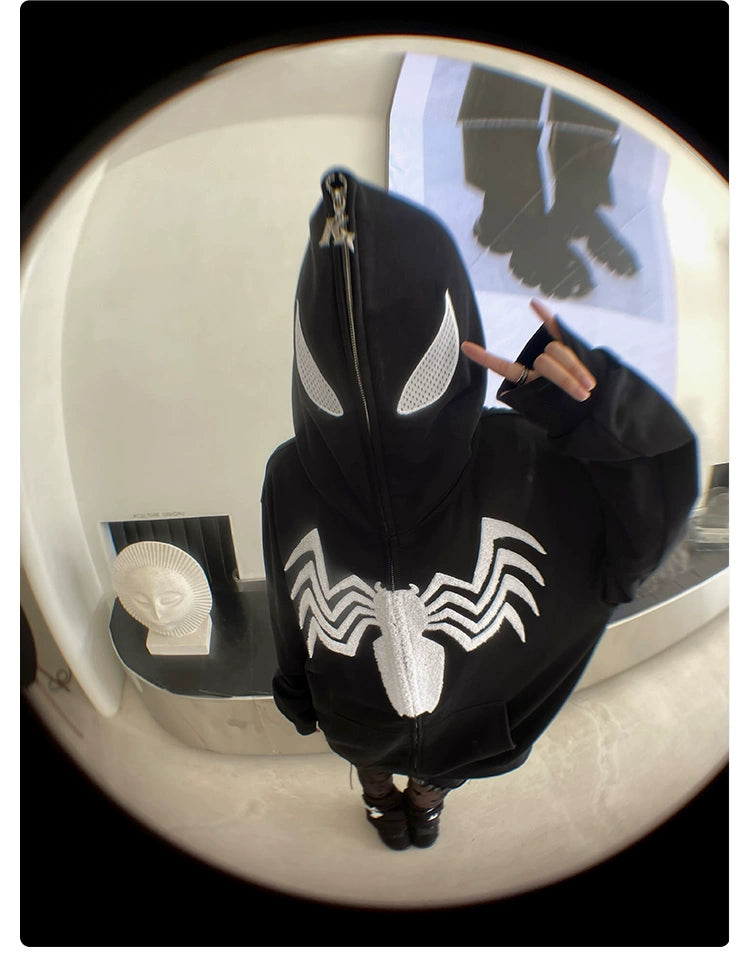 A person in a Seakoff Venom Spider Hoodie, an oversized black and white zip-up featuring a white spider emblem and eye designs, zips it up over the face while making a peace sign. A fisheye lens captures a white sculpture head and artwork in the background, perfect for streetwear fans.