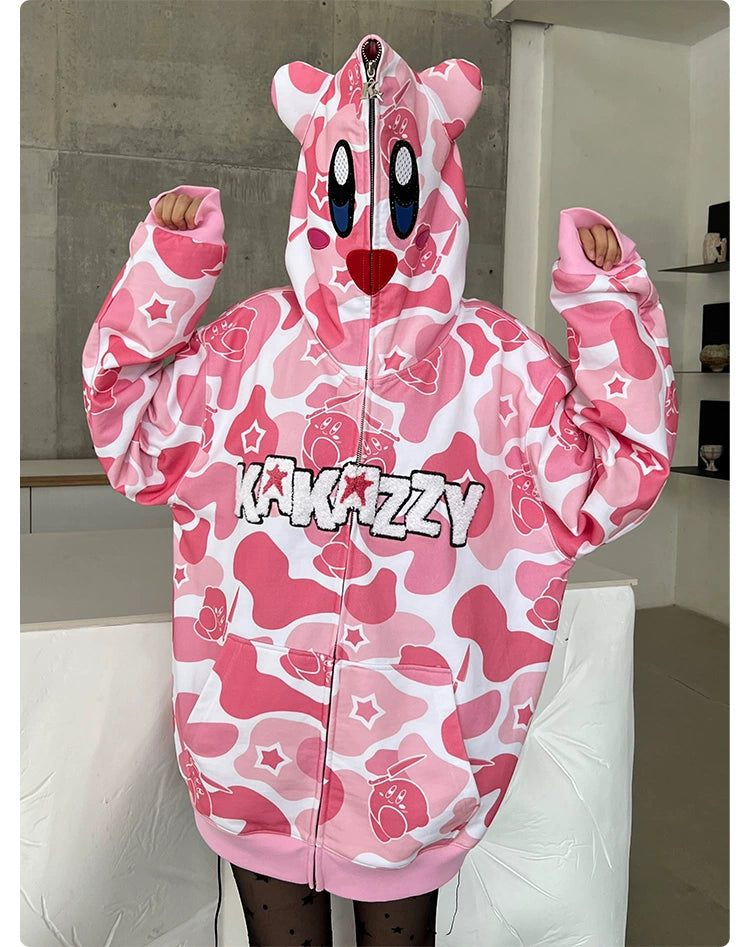A person wears a Seakoff Pink Kirby-Inspired Hoodie, featuring pink and white camo, cartoon eyes, and 3D ears on the hood. "KAKAZZY" is emblazoned on the front as they raise their hands in a minimalistic room with a concrete wall and shelf.