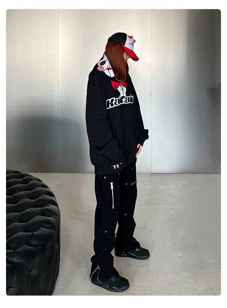Indoors, a person dons a Seakoff Saw-Inspired Hoodie—a 400GSM oversized black zip-up horror movie sweatshirt—paired with a red cap and zipper-detailed black pants. The minimalist room has a dark tufted seat on the left, with light-colored flooring and plain walls enhancing an eerie vibe.