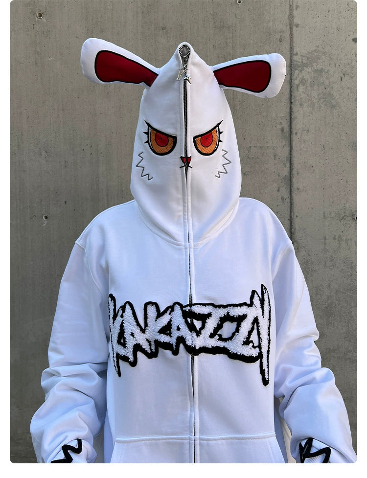 Someone is wearing the Seakoff Fierce Bunny Hoodie - a statement piece with an oversized fit, featuring a red-eyed bunny ear design and embroidered face on the hood. Bold black text adorns the front against a simplistic gray concrete wall, enhancing its striking appearance.