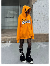 A person wearing a Seakoff Halloween Pumpkin Hoodie—an oversized, 400GSM orange zip-up sweatshirt adorned with Halloween graphics—paired with black star-patterned tights and black leg warmers featuring white skulls, stands on a sidewalk against a gray background.