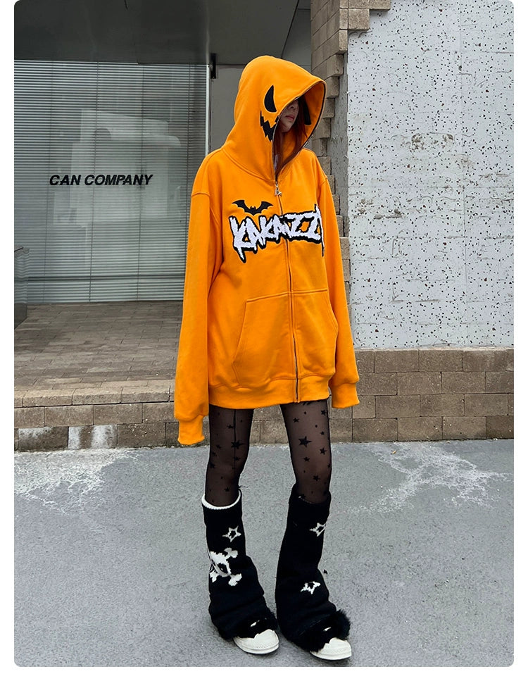 A person wearing a Seakoff Halloween Pumpkin Hoodie—an oversized, 400GSM orange zip-up sweatshirt adorned with Halloween graphics—paired with black star-patterned tights and black leg warmers featuring white skulls, stands on a sidewalk against a gray background.