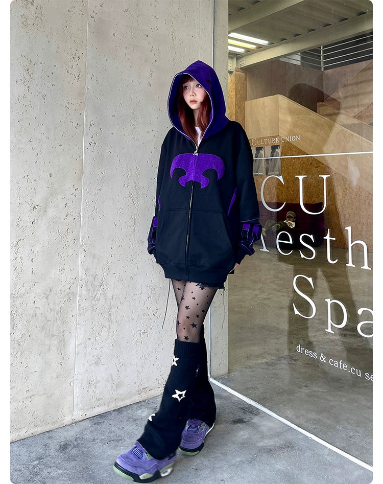 A person leans against a wall wearing the Seakoff Dark Hero Hoodie, an oversized black and purple superhero zip-up sweatshirt, complemented by star-patterned tights and purple sneakers. A glass window with partially visible text in the background enhances the superhero streetwear vibe.
