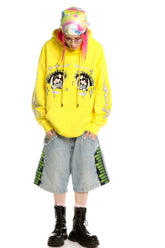 A streetwear lover wears a Seakoff Kawaii Aesthetic Crying Eyes Hoodie featuring sleeve graphics. Paired with baggy denim shorts and black boots, their pink hair peeks from under a multicolored beanie. Glasses against the white background enhance the anime-inspired charm.