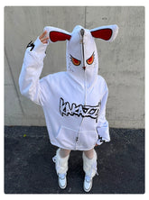 A person stands against a concrete wall wearing the Fierce Bunny Hoodie by Seakoff. This 400GSM cotton hoodie features red eyes and rabbit ears on the hood with a stylized front print and an oversized fit. They pair this with furry white boots, complementing the dark pavement beneath.