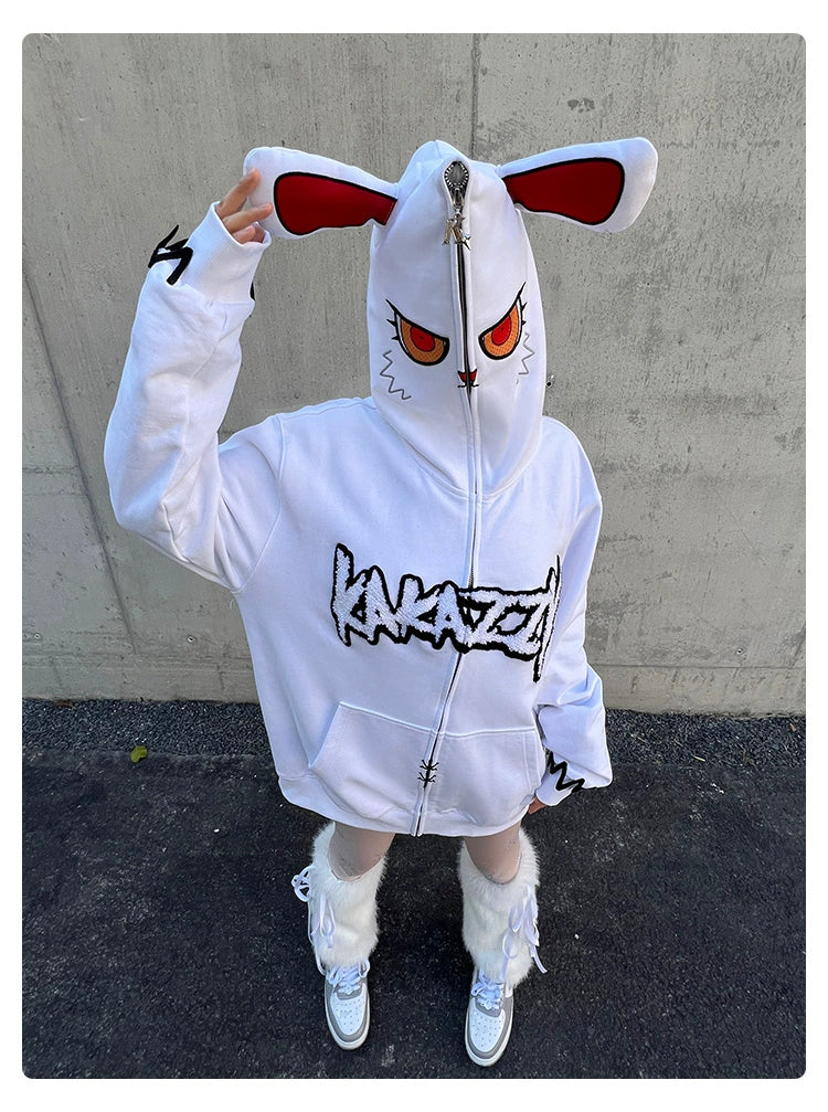 A person stands against a concrete wall wearing the Fierce Bunny Hoodie by Seakoff. This 400GSM cotton hoodie features red eyes and rabbit ears on the hood with a stylized front print and an oversized fit. They pair this with furry white boots, complementing the dark pavement beneath.