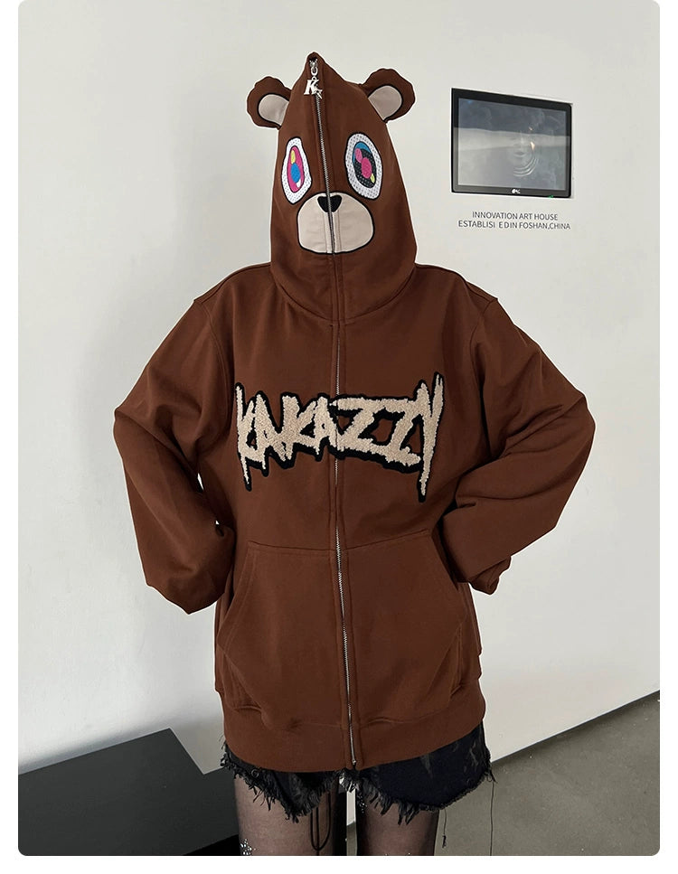 A person in a Seakoff Cute Bear Hoodie, an oversized brown 400GSM streetwear sweatshirt featuring ears and a zip-up design, confidently stands against a white wall. With their hands in pockets, the hoodie fully covers their head, merging comfort with urban style.