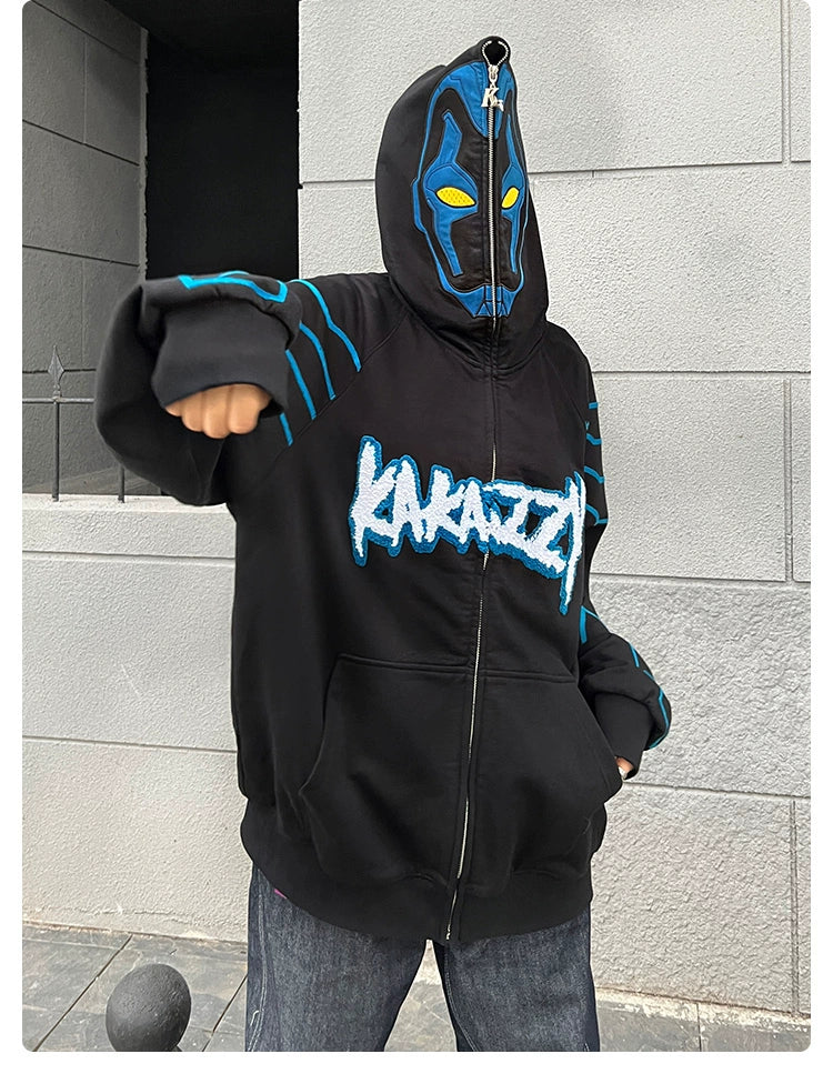 A person wearing a Seakoff Cyberpunk Superhero-Inspired Hoodie, a 400GSM oversized black zip-up sci-fi sweatshirt with blue designs and "KAKAZA" text, sports a matching mask while striking a stylish pose against a gray brick wall.