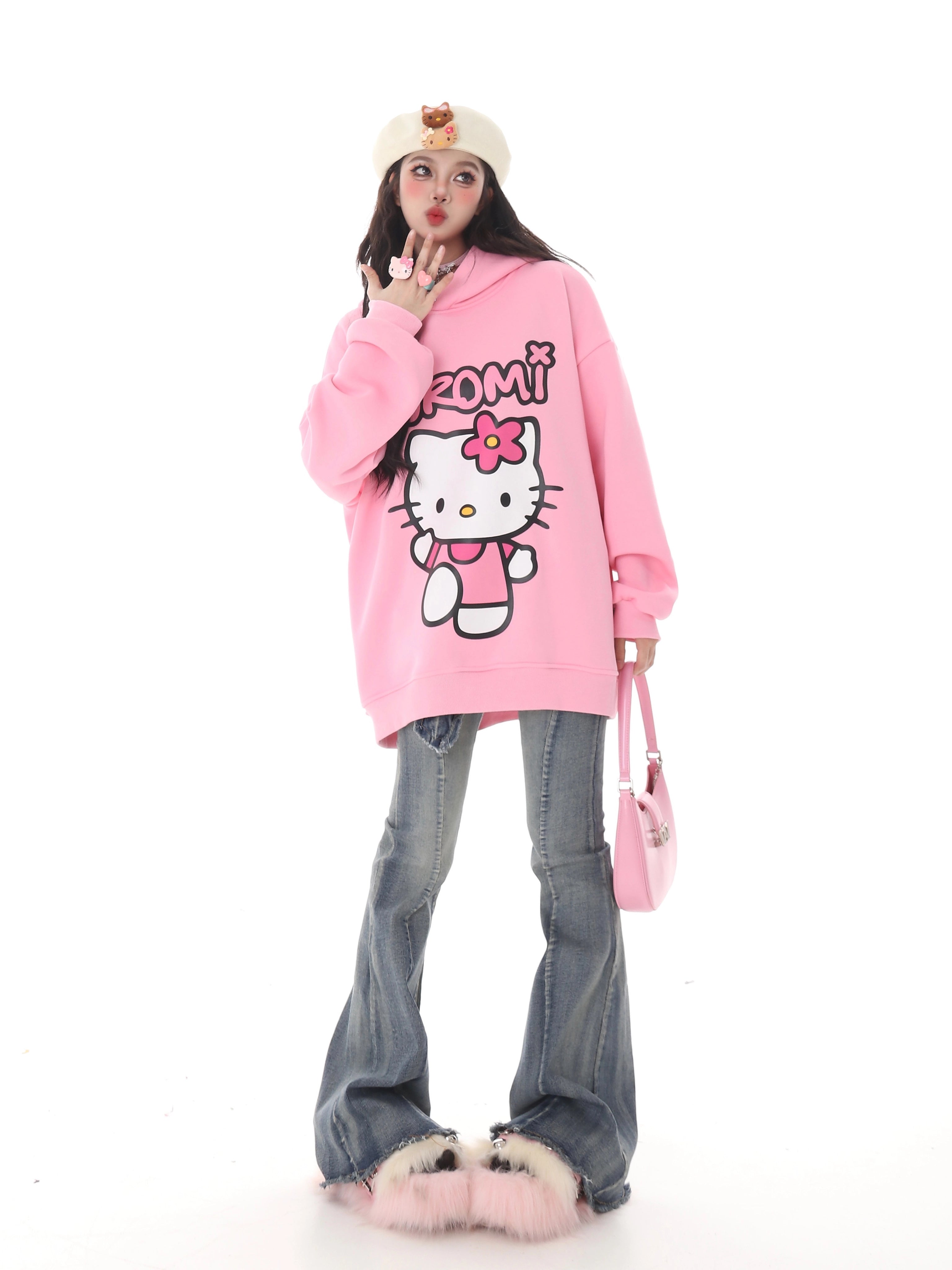 A person in a pink Seakoff Kawaii Hello Kitty oversized hoodie and flared jeans poses against a white background, showcasing kawaii fashion. They accessorize with a white beret, pink handbag, and fluffy shoes. Sanrio enthusiasts will love their playful gesture with fingers near their mouth.
