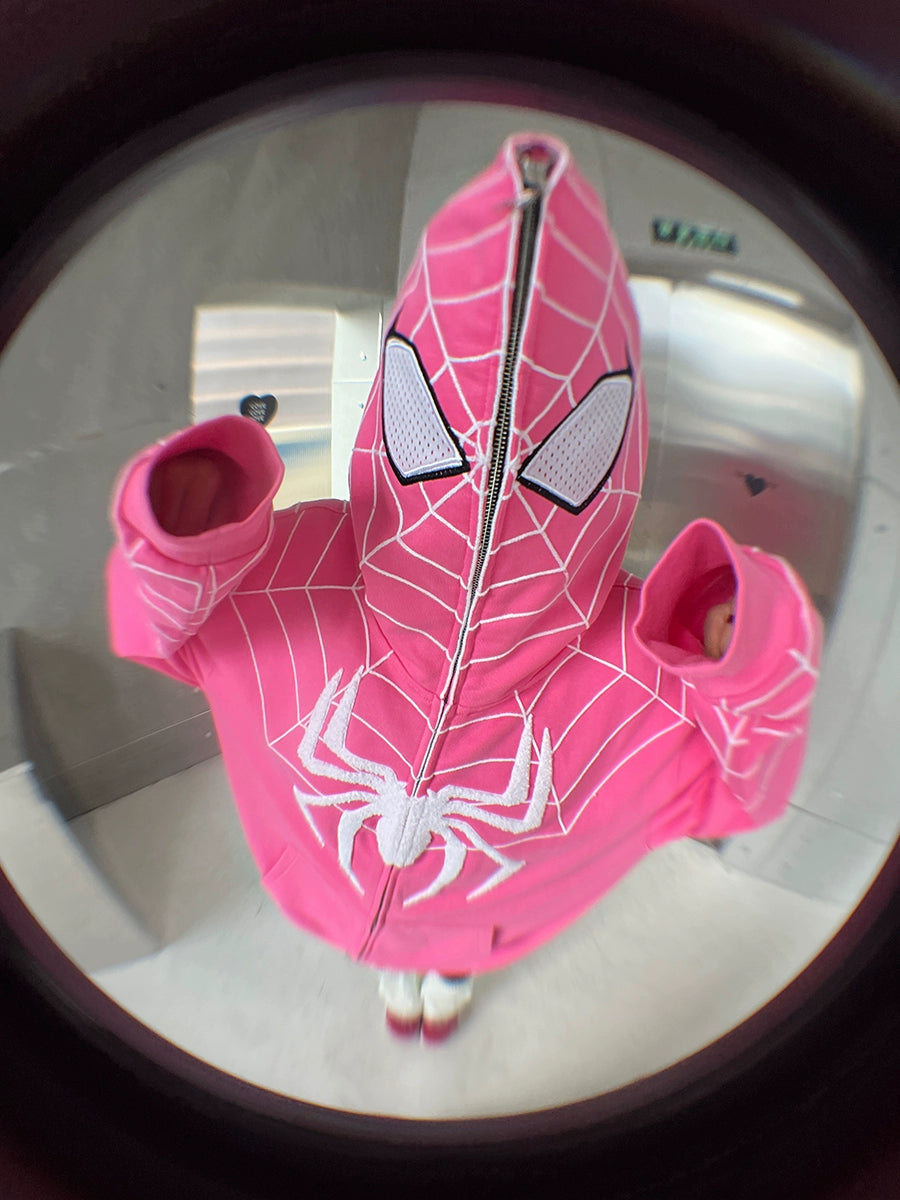 A child poses indoors wearing a Seakoff Spider-Inspired Hoodie, an oversized pink zip-up sweatshirt with a web design and large white spider logo. Captured with a fisheye lens, it&