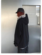A person with long red hair and a black cap is wearing the Spirited Away No-Face Anime Hoodie, appealing to anime fans. They stand facing a metallic wall labeled "CULTURE-UNION," capturing the essence of Spirited Away in their unique style.