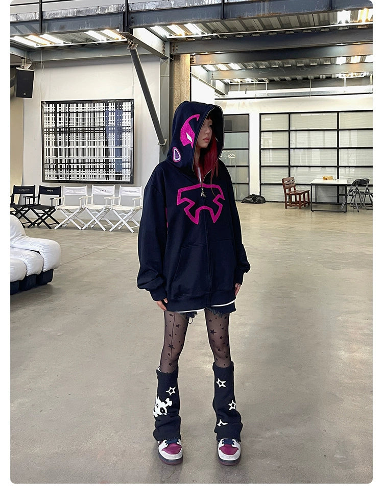A person in a Seakoff Futuristic Warrior Hoodie—a 400GSM oversized sci-fi pink and black zip-up sweatshirt—pairs it with a plaid skirt, star-patterned sheer tights, and tall black leg warmers with star patches. They stand in a spacious room with industrial decor, visible chairs, and large artwork.