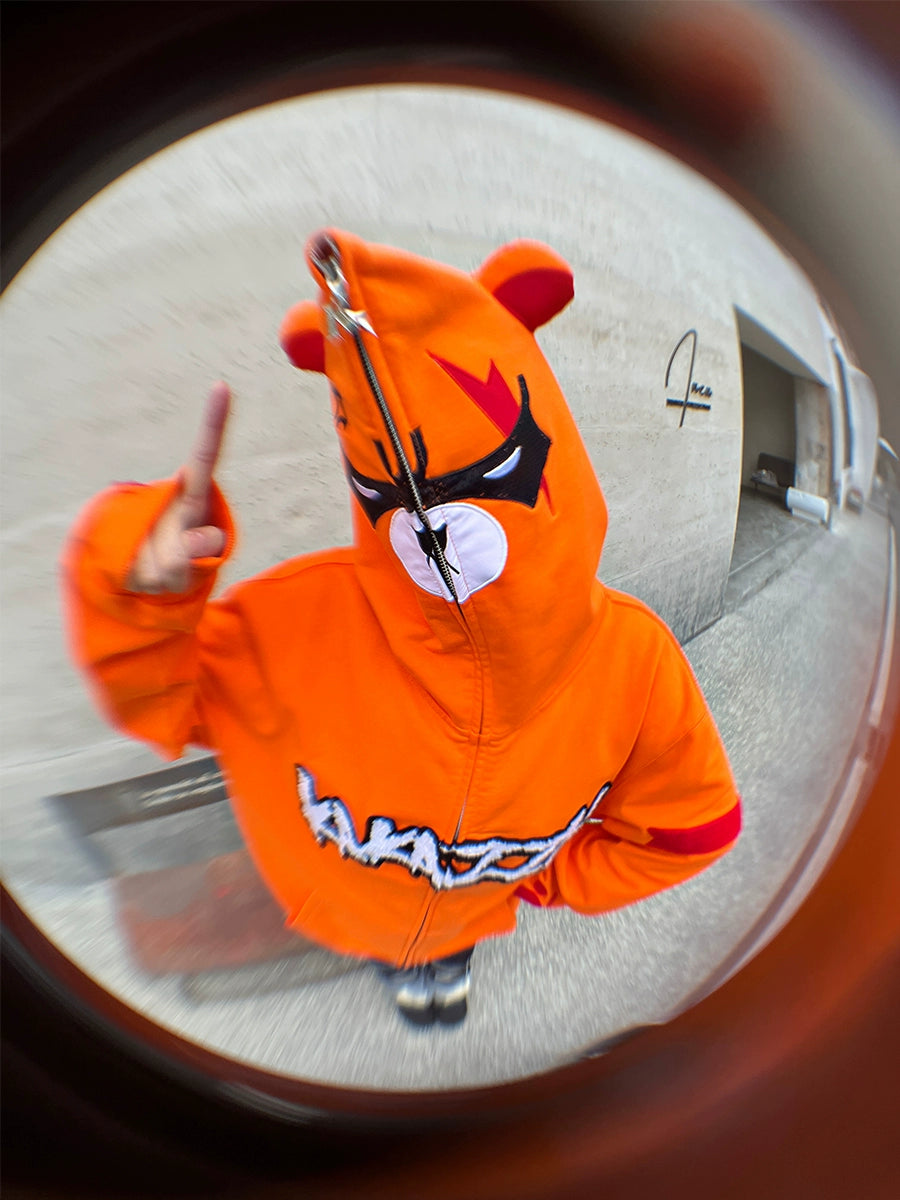 A person wearing a Seakoff Furious Orange Bear Hoodie with an angry bear design and red embroidery, face hidden by the fully zipped hood, gestures a peace sign. The concrete backdrop is captured with a fisheye effect, showcasing the premium 400GSM cotton&