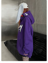 A person in a Seakoff Fun Cartoon Face Hoodie, an oversized 400GSM purple zip-up streetwear sweatshirt, stands by a black couch. They have long hair and wear a gray cap with ear-like extensions. The concrete and metallic walls backdrop enhances the streetwear vibe.