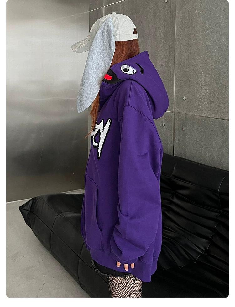 A person in a Seakoff Fun Cartoon Face Hoodie, an oversized 400GSM purple zip-up streetwear sweatshirt, stands by a black couch. They have long hair and wear a gray cap with ear-like extensions. The concrete and metallic walls backdrop enhances the streetwear vibe.