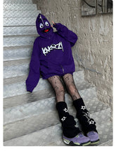 A person sits on metal stairs, wearing a Seakoff Fun Cartoon Face Hoodie—an oversized 400GSM purple zip-up streetwear sweatshirt—with patterned fishnet tights and black leg warmers featuring stars. Their sneakers are purple and green, while the peeling paint wall enhances the streetwear vibe.