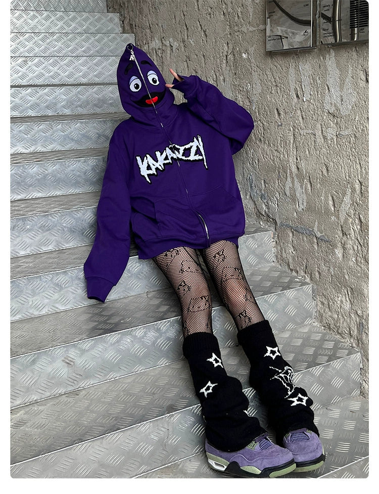 A person sits on metal stairs, wearing a Seakoff Fun Cartoon Face Hoodie—an oversized 400GSM purple zip-up streetwear sweatshirt—with patterned fishnet tights and black leg warmers featuring stars. Their sneakers are purple and green, while the peeling paint wall enhances the streetwear vibe.