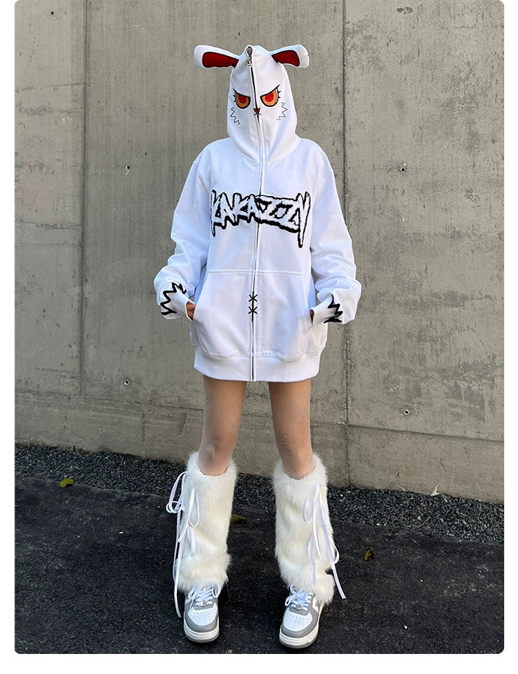 A person in a Seakoff Fierce Bunny Hoodie featuring red eyes and rabbit ear details stands against a concrete wall. The oversized 400GSM cotton hoodie is matched with white furry leg warmers and shoes, while they maintain a neutral expression.