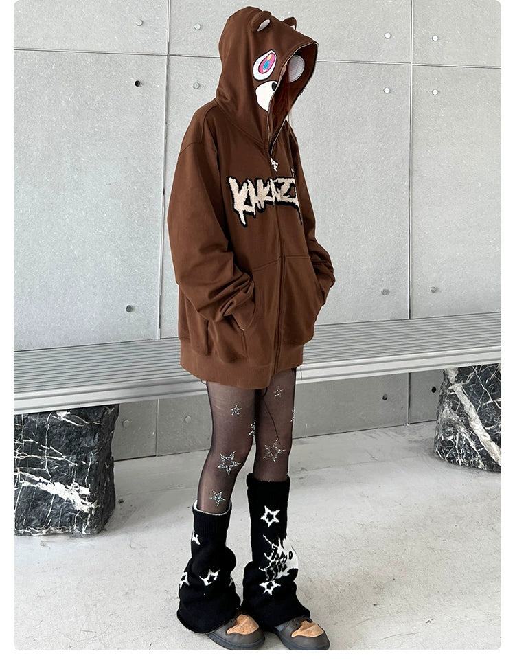 A person wears a Seakoff Cute Bear Hoodie, an oversized brown 400GSM zip-up streetwear sweatshirt with ears, cartoon eyes on the hood, sheer black star-patterned tights, black fuzzy leg warmers, and sandals against a gray wall and marble bench in the background.