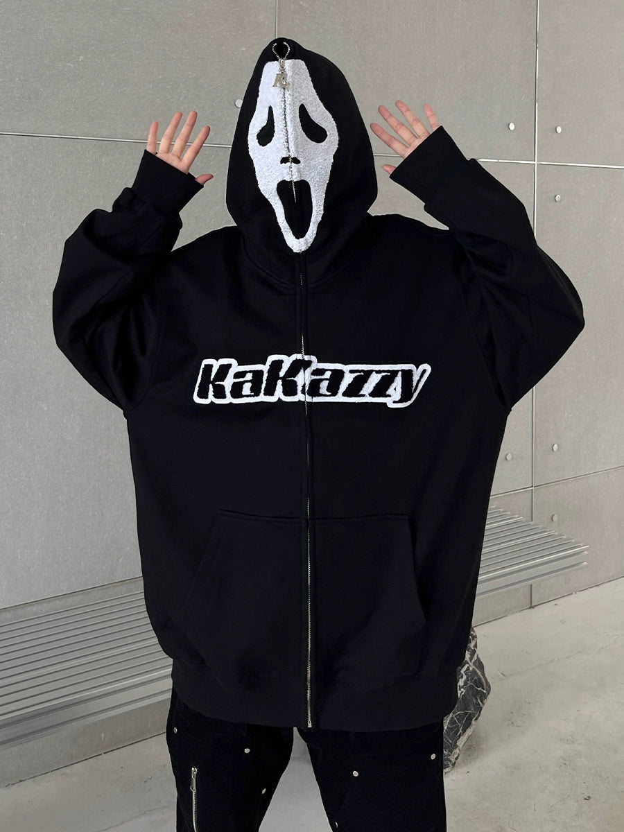 A person stands with hands raised, wearing the Seakoff Scream-Inspired Hoodie, a 400GSM oversized black zip-up sweatshirt featuring subtle Ghostface mask designs. The textured gray wall and a metal bench in the background perfectly complement this streetwear look.