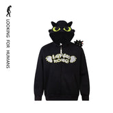 The Seakoff Cute Night Fury Dragon Hoodie is a black anime-inspired full zip hoodie featuring large green eyes and 3D ears on the hood, with "Looking for Humans" on the chest. Ideal for fans, it includes a small plush toy resembling Night Fury from How to Train Your Dragon on the shoulder.
