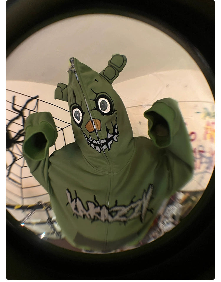A person wears an oversized green Seakoff Springtrap-Inspired Hoodie, showcasing a cartoonish face and ears on the hood with a zipper-mouth design. The sleeves are up, against graffiti and metal bars. The fisheye lens effect enhances the eerie vibe for Five Nights at Freddy&