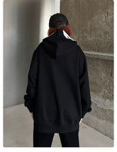 A person with long hair, wearing a Seakoff Scream-Inspired Hoodie—an oversized black zip-up horror movie sweatshirt—faces a concrete wall. Their back is turned towards the camera in a minimalist indoor setting—perfect for horror fans envisioning lurking shadows.