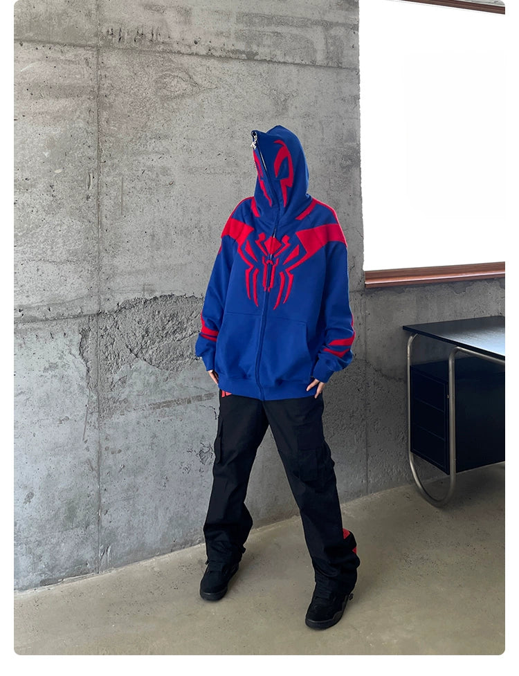 A person stands indoors on a concrete floor, wearing the Superhero Spider Hoodie by Seakoff. This oversized blue and red zip-up sweatshirt features a red spider emblem. They have their hood up, and black pants complete the outfit. A desk is partially visible on the right side.