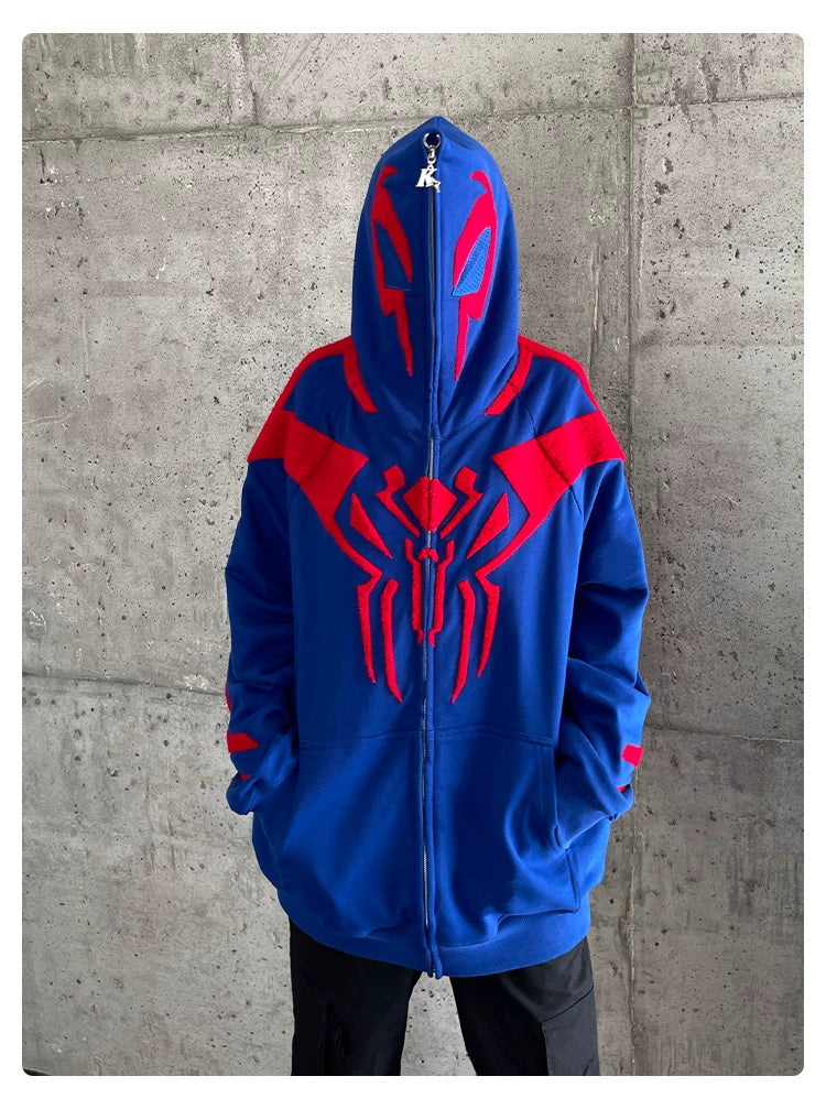 A person in a Seakoff Superhero Spider Hoodie, a blue and red oversized zip-up sweatshirt for fans, stands against a gray concrete wall. The hood is up, concealing their face as they casually keep their hands in their pockets.