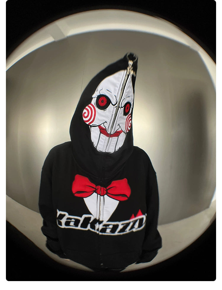 A person wears a Seakoff Saw-Inspired hoodie, zipped over their face, revealing a design reminiscent of Billy the Puppet with spirals and graphics of a red bow tie and "Kazazu." Captured with a fisheye lens, this 400GSM oversized black zip-up appeals to horror movie enthusiasts.