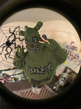 Wearing the oversized Springtrap-Inspired Hoodie by Seakoff, a green 400GSM zip-up with cartoon graphics and fabric ears, they hide their face. Spider decorations and graffiti surround them—a perfect scene for Five Nights at Freddy&
