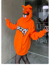 A person poses playfully with arms outstretched in front of a building, wearing an oversized Seakoff Furious Orange Bear Hoodie featuring an angry bear face on the hood and red "MADVERY" embroidery. They pair it with star-patterned tights.