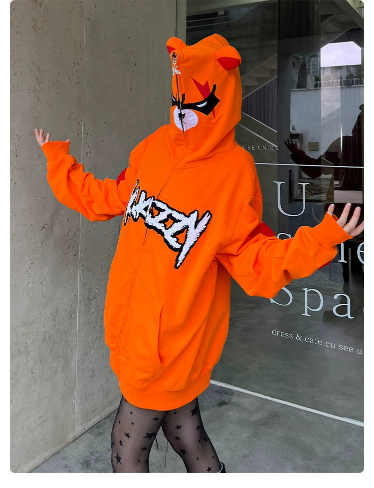 A person poses playfully with arms outstretched in front of a building, wearing an oversized Seakoff Furious Orange Bear Hoodie featuring an angry bear face on the hood and red "MADVERY" embroidery. They pair it with star-patterned tights.