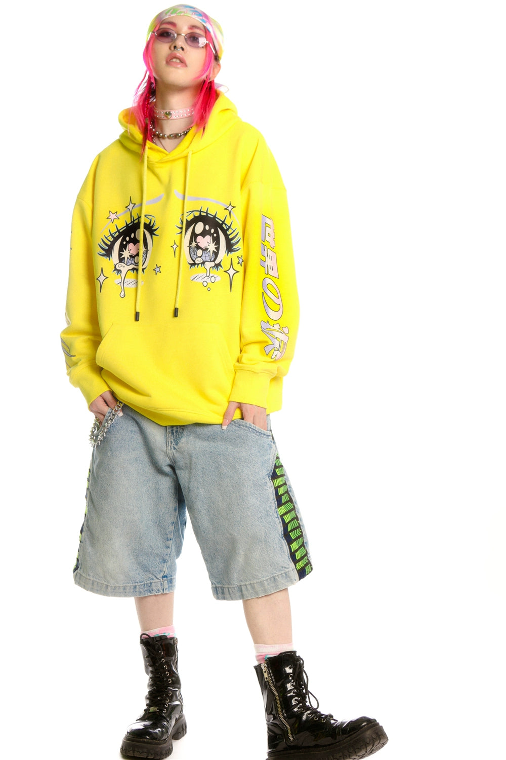 A person with pink hair, glasses, and a colorful headband wears a Seakoff Kawaii Aesthetic Crying Eyes Hoodie—Pastel & Dark Anime Pullover with Sleeve Graphics. They pair their look with denim shorts, black boots, a choker necklace, and bracelets against a white background.