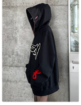 A person wearing a Berserk Griffith Dark Knight Hoodie stands sideways near a concrete wall, facing a glass door. The oversized 400GSM black zip-up streetwear features a red emblem on the arm and graphic design on the front, with the hood up obscuring their face.
