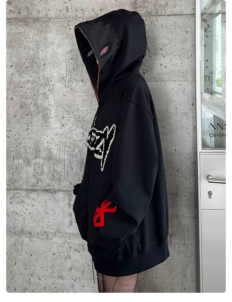 A person wearing a Berserk Griffith Dark Knight Hoodie stands sideways near a concrete wall, facing a glass door. The oversized 400GSM black zip-up streetwear features a red emblem on the arm and graphic design on the front, with the hood up obscuring their face.