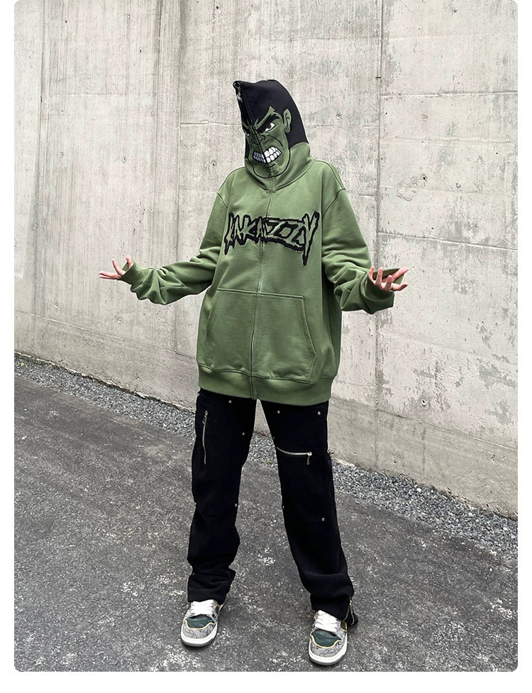 A streetwear enthusiast poses on a paved surface before a concrete wall, wearing a Seakoff Hulk-Inspired Hoodie - 400GSM Oversized Green Zip-Up Superhero Sweatshirt with black pants. The hood is up, and their hands are open and slightly raised.