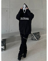 A person wearing a Seakoff Scream-Inspired Hoodie—an oversized black zip-up horror movie sweatshirt—stands by a concrete wall, adjusting their Ghostface mask. Paired with black pants and shoes, it’s a look any horror fan would appreciate. In the background are a metal bench and abstract stone base.
