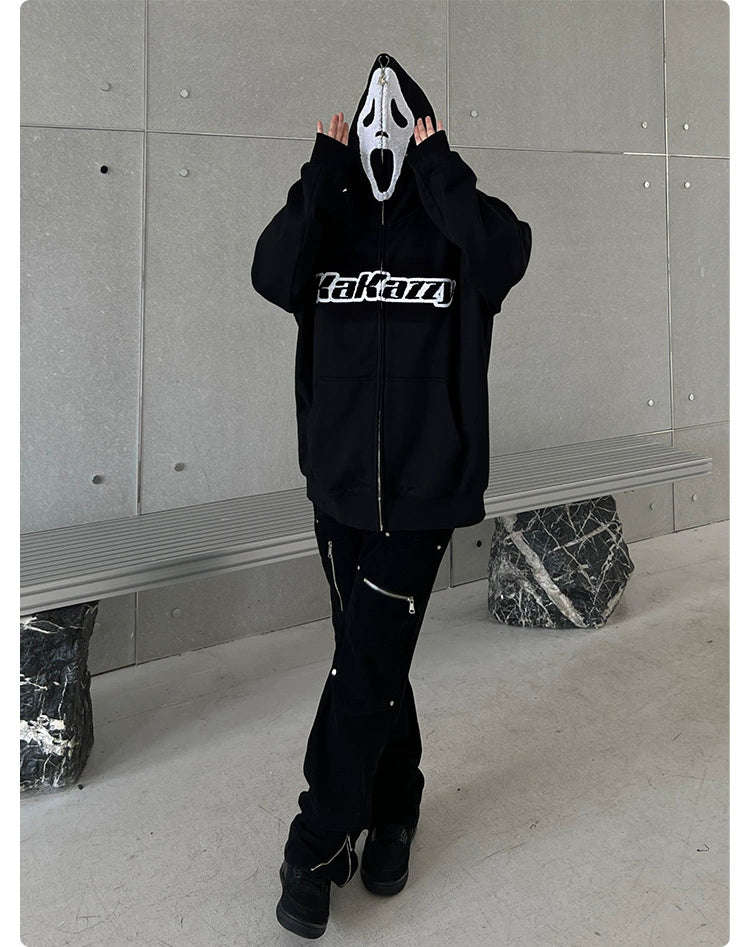 A person wearing a Seakoff Scream-Inspired Hoodie—an oversized black zip-up horror movie sweatshirt—stands by a concrete wall, adjusting their Ghostface mask. Paired with black pants and shoes, it’s a look any horror fan would appreciate. In the background are a metal bench and abstract stone base.