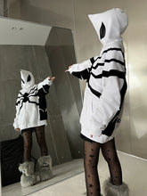 In a grey-walled room, someone stands before a mirror, their face hidden by the hood featuring a spider emblem from Seakoff&