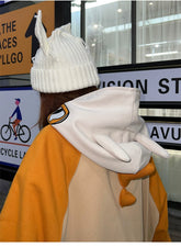 Wearing a whimsical white knit hat with bunny ears and the Pokémon Cubone Full Zip hoodie, inspired by nostalgic anime cosplay with charming ear details, this individual showcases playful fashion against a blurred sign and cyclist backdrop.