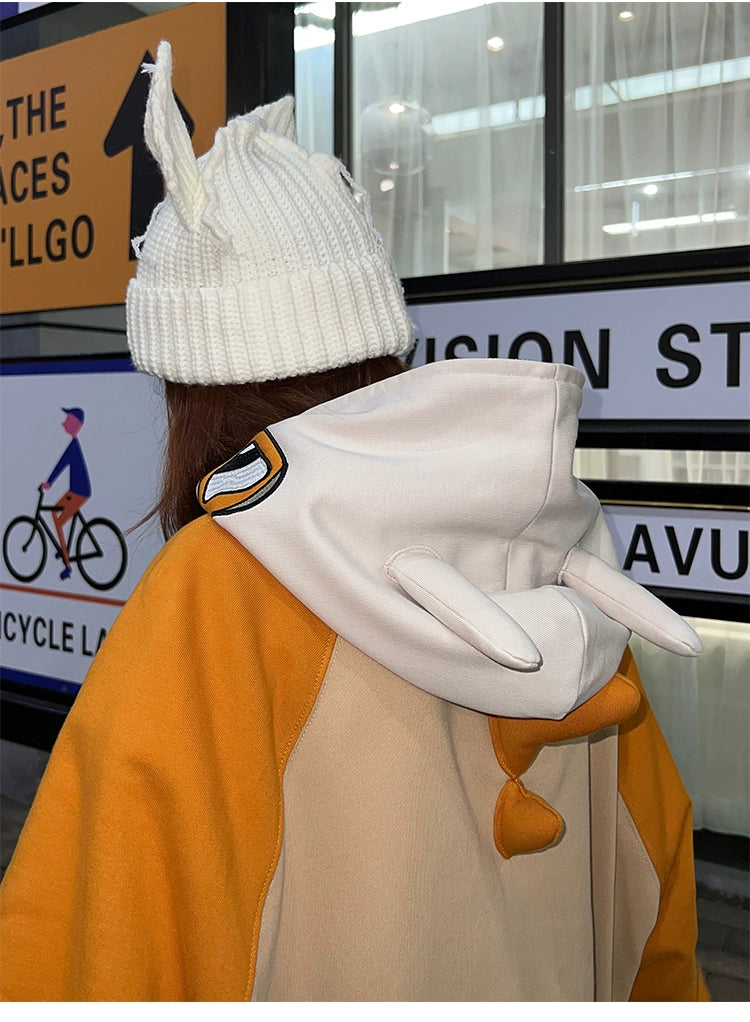 Wearing a whimsical white knit hat with bunny ears and the Pokémon Cubone Full Zip hoodie, inspired by nostalgic anime cosplay with charming ear details, this individual showcases playful fashion against a blurred sign and cyclist backdrop.