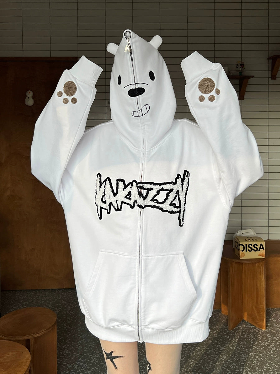 Indoors, someone models the Seakoff Cute White Bear Hoodie. This oversized 400GSM hoodie features bear ears and paw print-patterned sleeves, highlighted by a polar bear design with black embroidery on the front. The room is accented with wooden furniture.