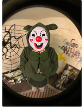 A person wearing a Seakoff Springtrap-Inspired Hoodie—a 400GSM oversized green zip-up sweatshirt—paired with a clown mask featuring red cheeks and a red nose stands indoors, surrounded by spiderweb decorations and graffiti, creating an ideal scene for Five Nights at Freddy&