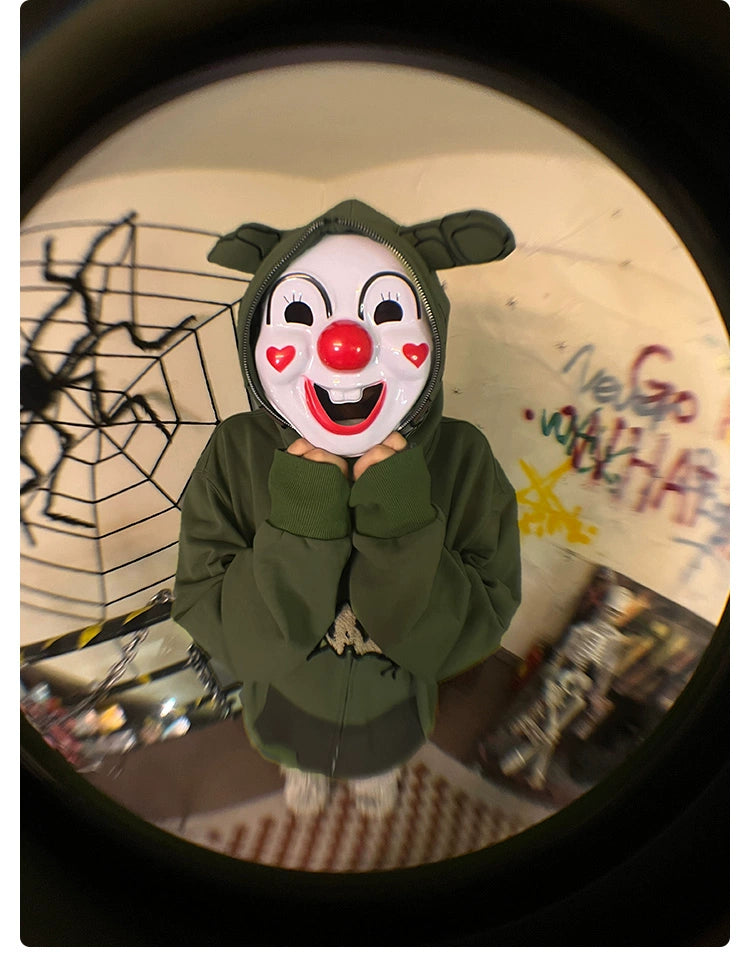 A person wearing a Seakoff Springtrap-Inspired Hoodie—a 400GSM oversized green zip-up sweatshirt—paired with a clown mask featuring red cheeks and a red nose stands indoors, surrounded by spiderweb decorations and graffiti, creating an ideal scene for Five Nights at Freddy&