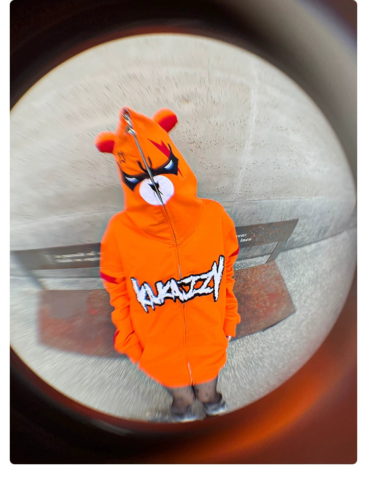 Wearing the Seakoff Furious Orange Bear Hoodie, featuring an Angry Bear cartoon face with ears and red embroidered graffiti-style text, a person stands against a concrete wall. The fisheye lens effect creates a circular distortion around them. The hoodie is made from premium 400GSM cotton for ultimate comfort.