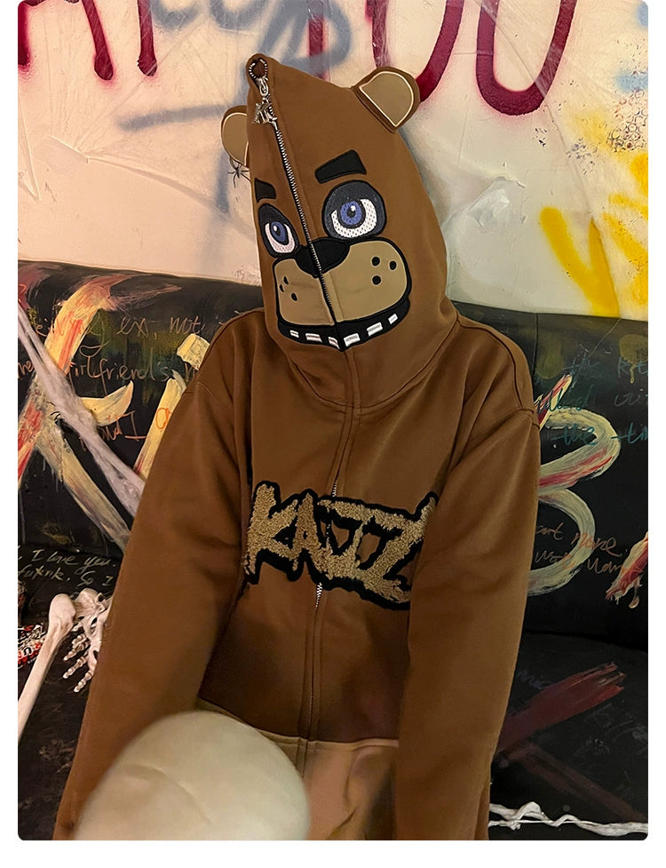 A person is wearing a Seakoff Freddy-Inspired Hoodie, showcasing a full-face design with eyes and snout. "KATSU" adorns the chest as they sit on a graffiti-covered sofa with a skeleton hand nearby—a perfect scene for Five Nights at Freddy&