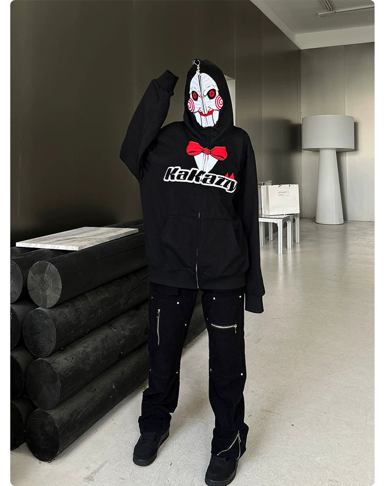 A person stands in a room wearing a Seakoff Saw-Inspired Hoodie, a 400GSM oversized black zip-up sweatshirt for horror movie fans. The mask, with colorful Billy the Puppet-like designs, is surrounded by a tall lamp, stacked black cylinders, and a white table with bags.