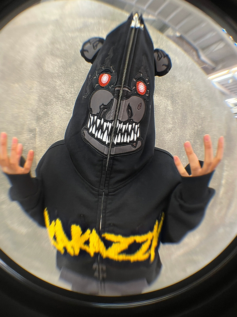 A horror fan sports a Seakoff Dark Bear Demon Hoodie, 400GSM cotton with a bear-shaped hood featuring red eyes and sharp teeth, and "YAZZY" embroidered in yellow. Their raised hands and the fisheye lens effect amplify the eerie vibe.