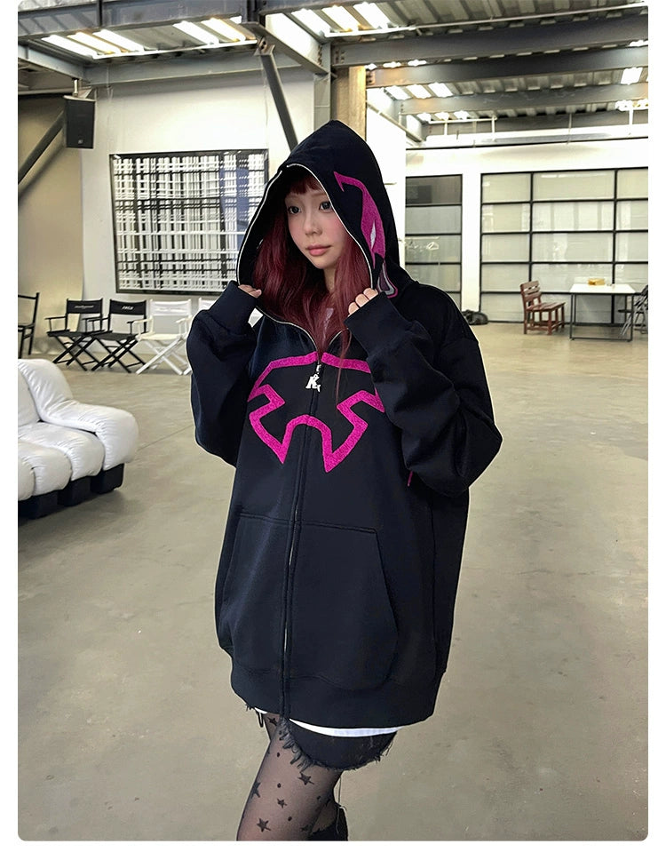 In a spacious, modern industrial room, someone with long hair wears a Seakoff Futuristic Warrior Hoodie—an oversized 400GSM sweatshirt in pink and black. With the hood up and camera-focused, they embody a sci-fi streetwear vibe enhanced by scattered furniture.
